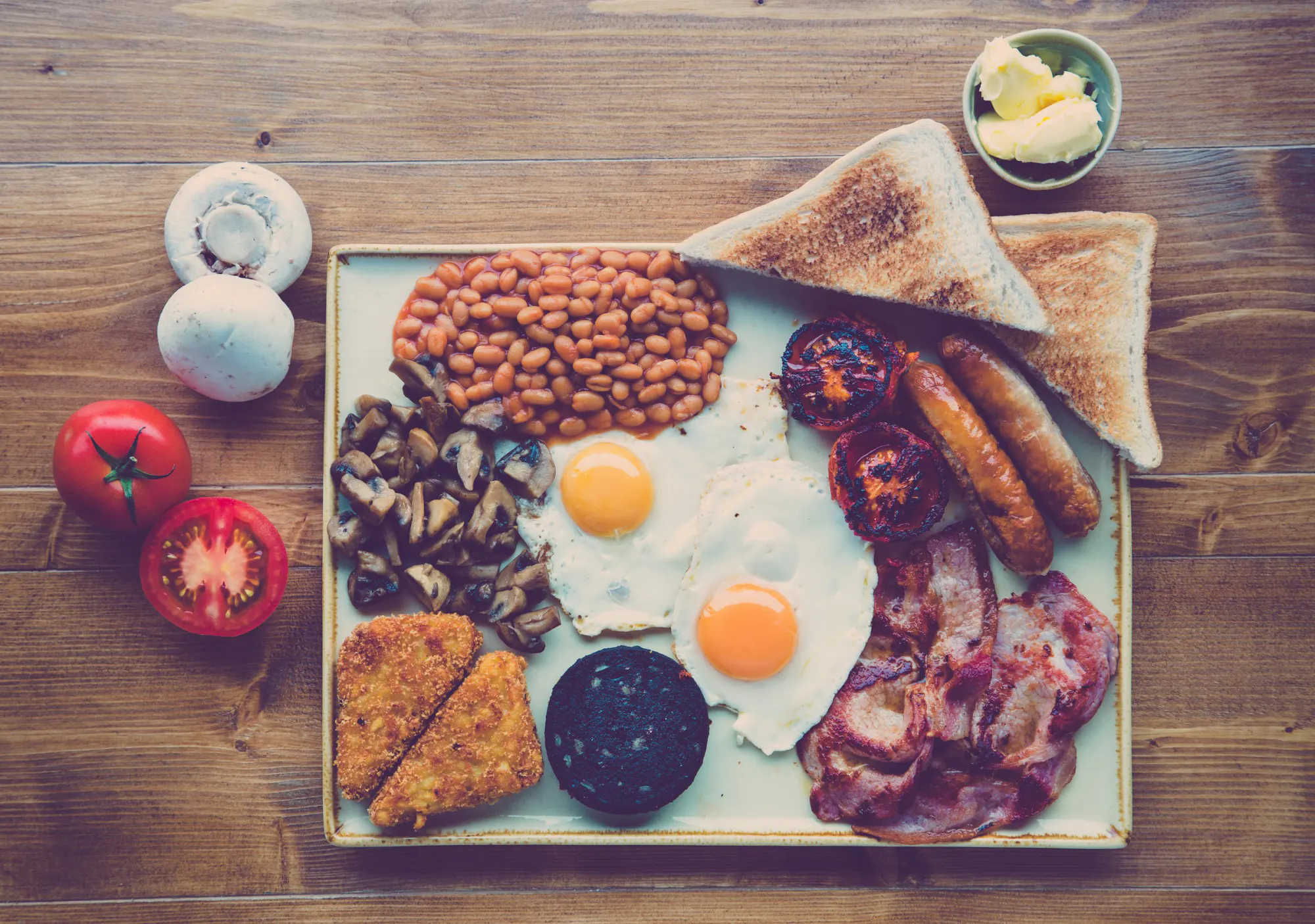 A full breakfast of sausages, bacon, fried eggs, toast, beans and tomatoes on a wooden table