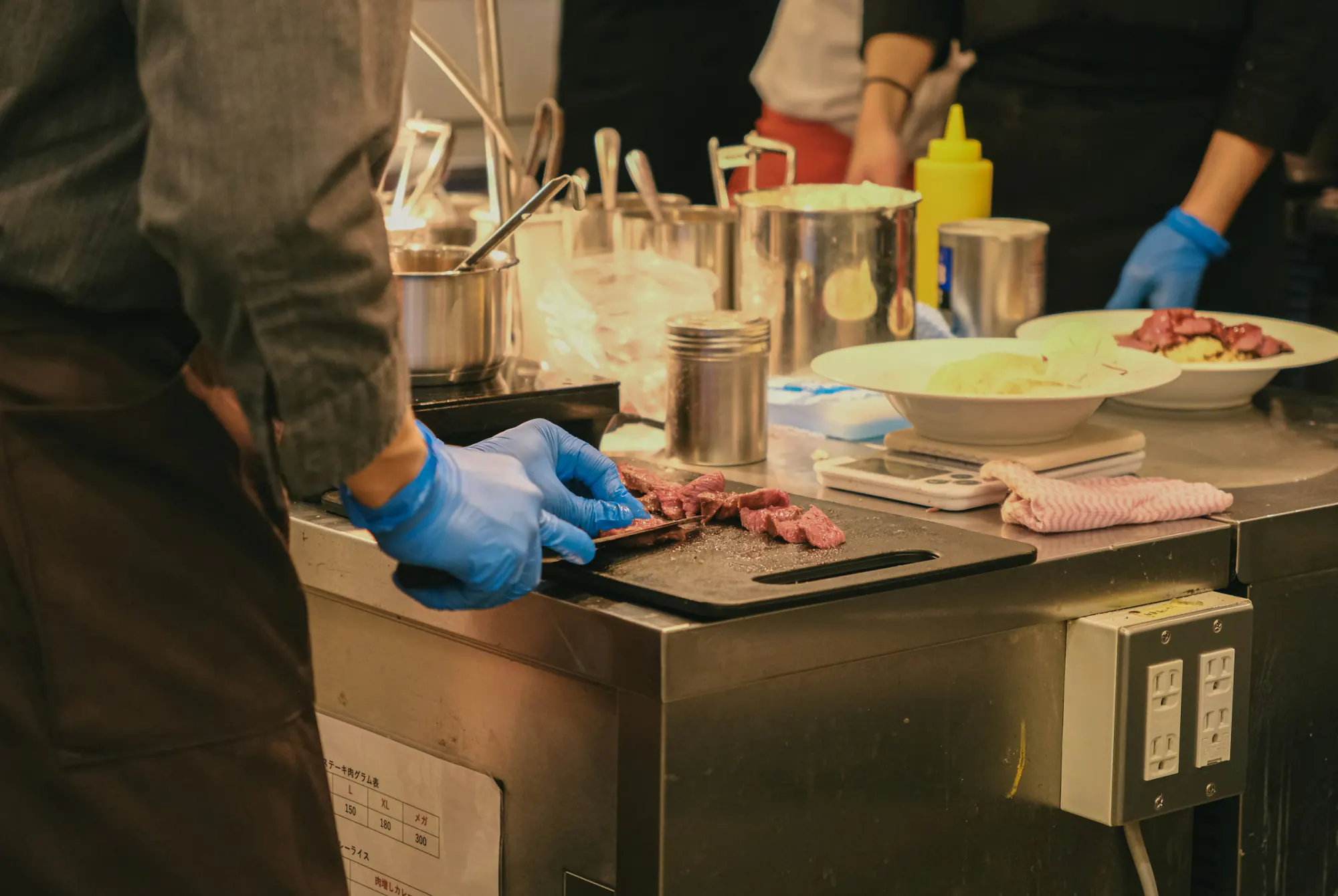 A cook in blue food-safety gloves preparing meat at a clean stainless-steel counter